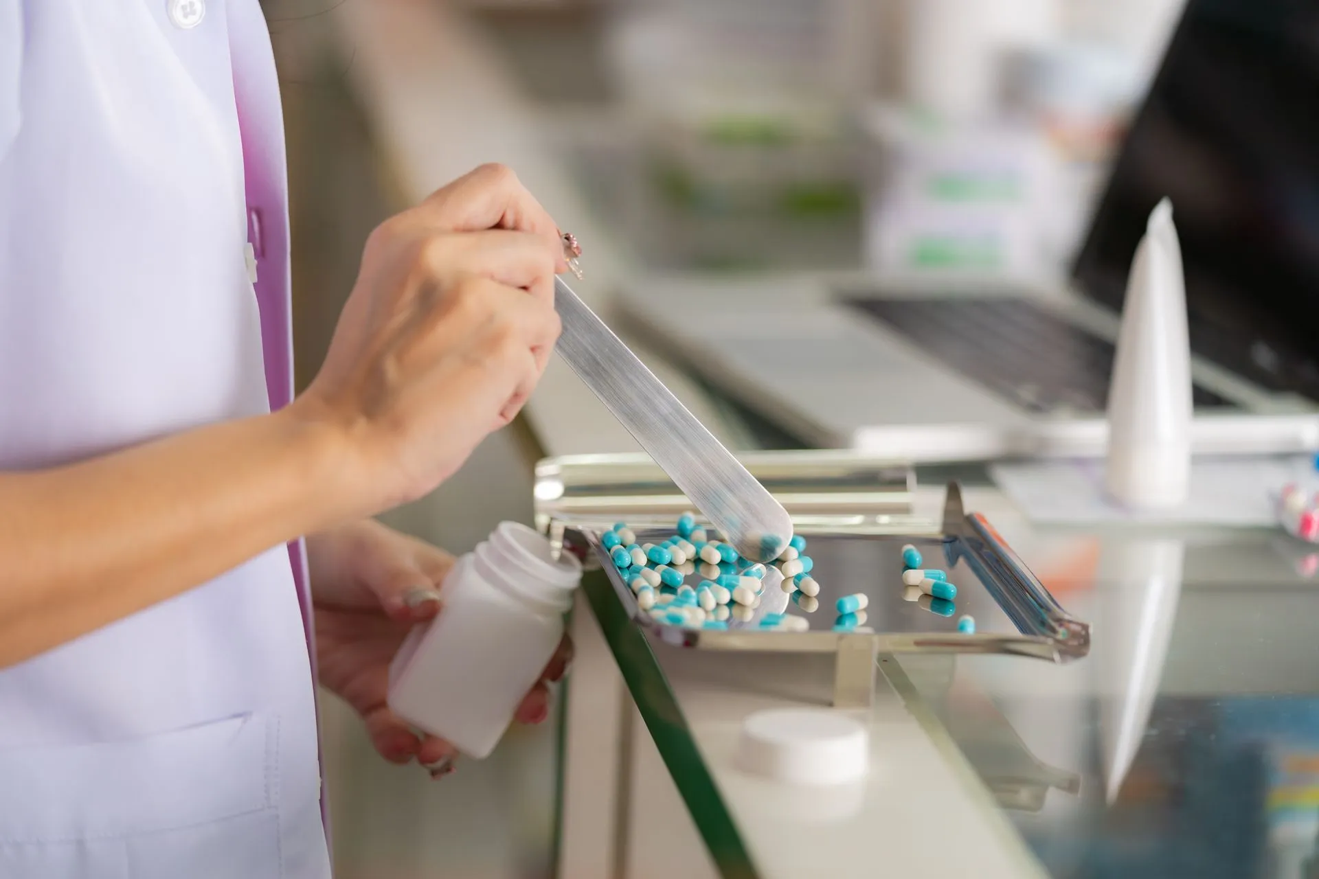 Pharmacist counting blue and white pills into a white prescription bottle at a pharmacy counter.