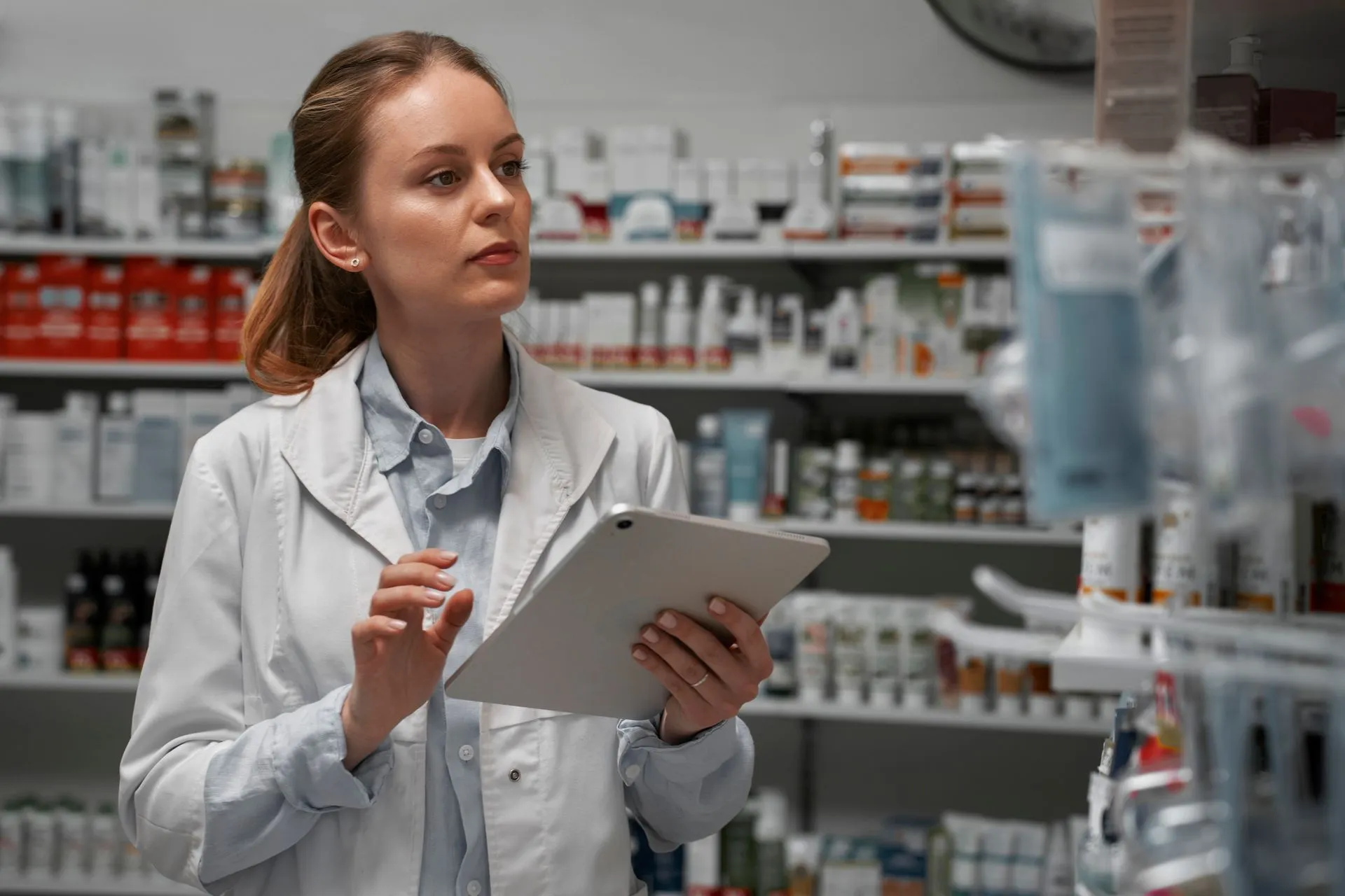 Pharmacist holding a tablet while checking stock on pharmacy shelves