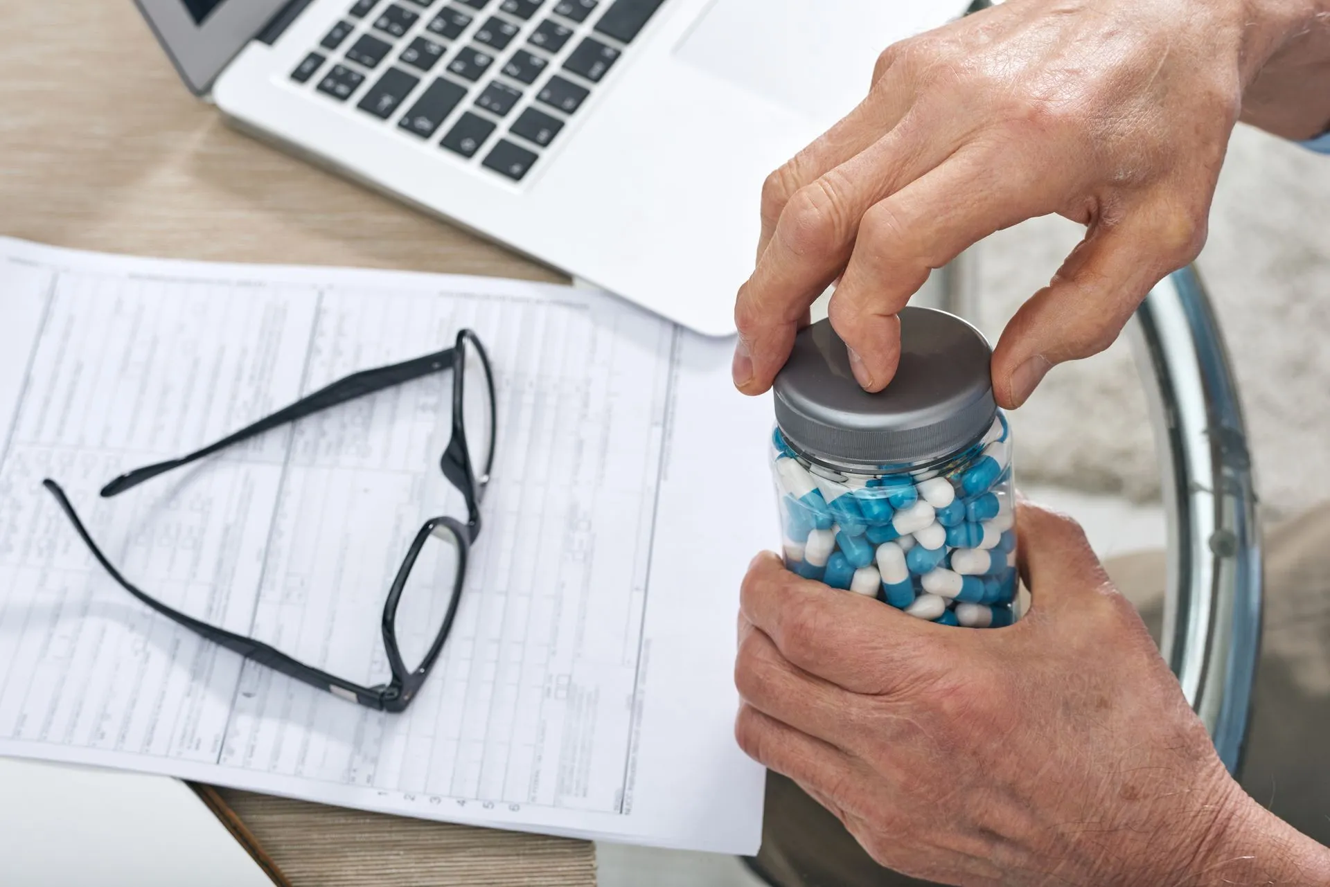 Senior man opening a jar of capsules at a desk with paperwork and reading glasses