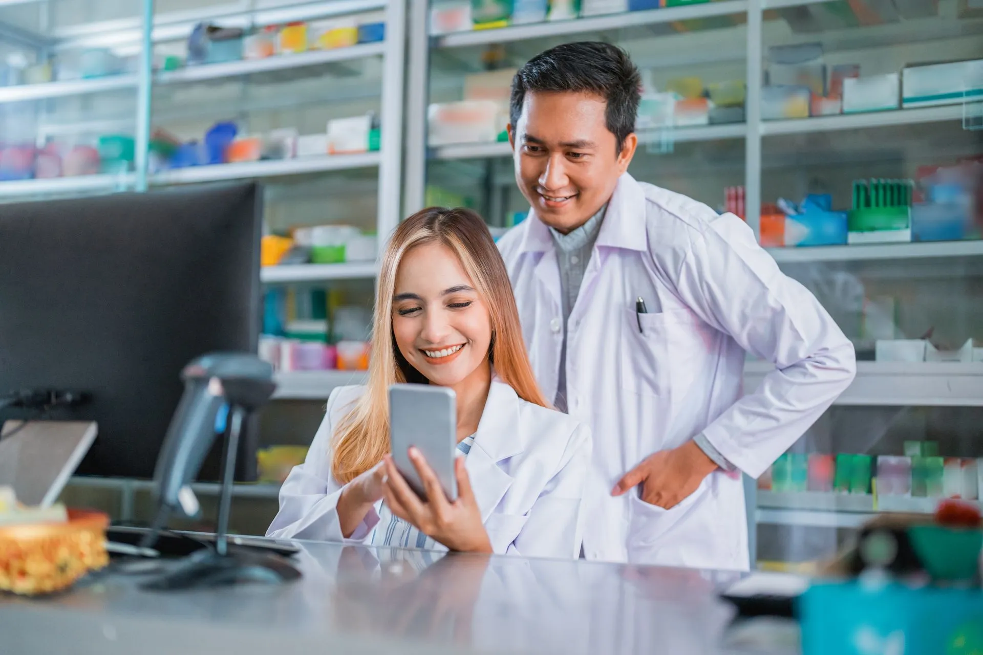Two smiling pharmacists looking at a smartphone together at the pharmacy counter