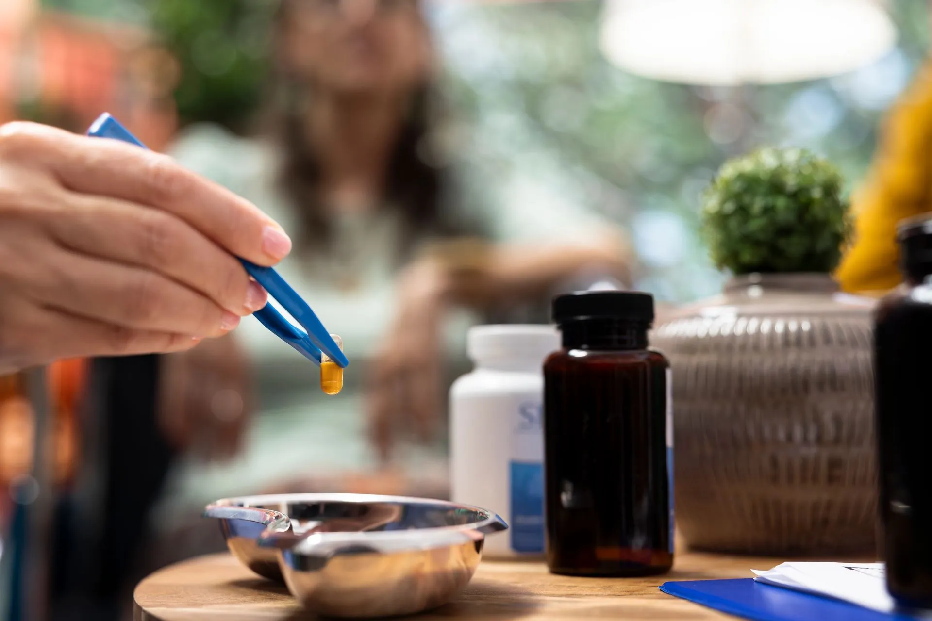 Hand using tweezers to pick up a softgel capsule beside supplement bottles