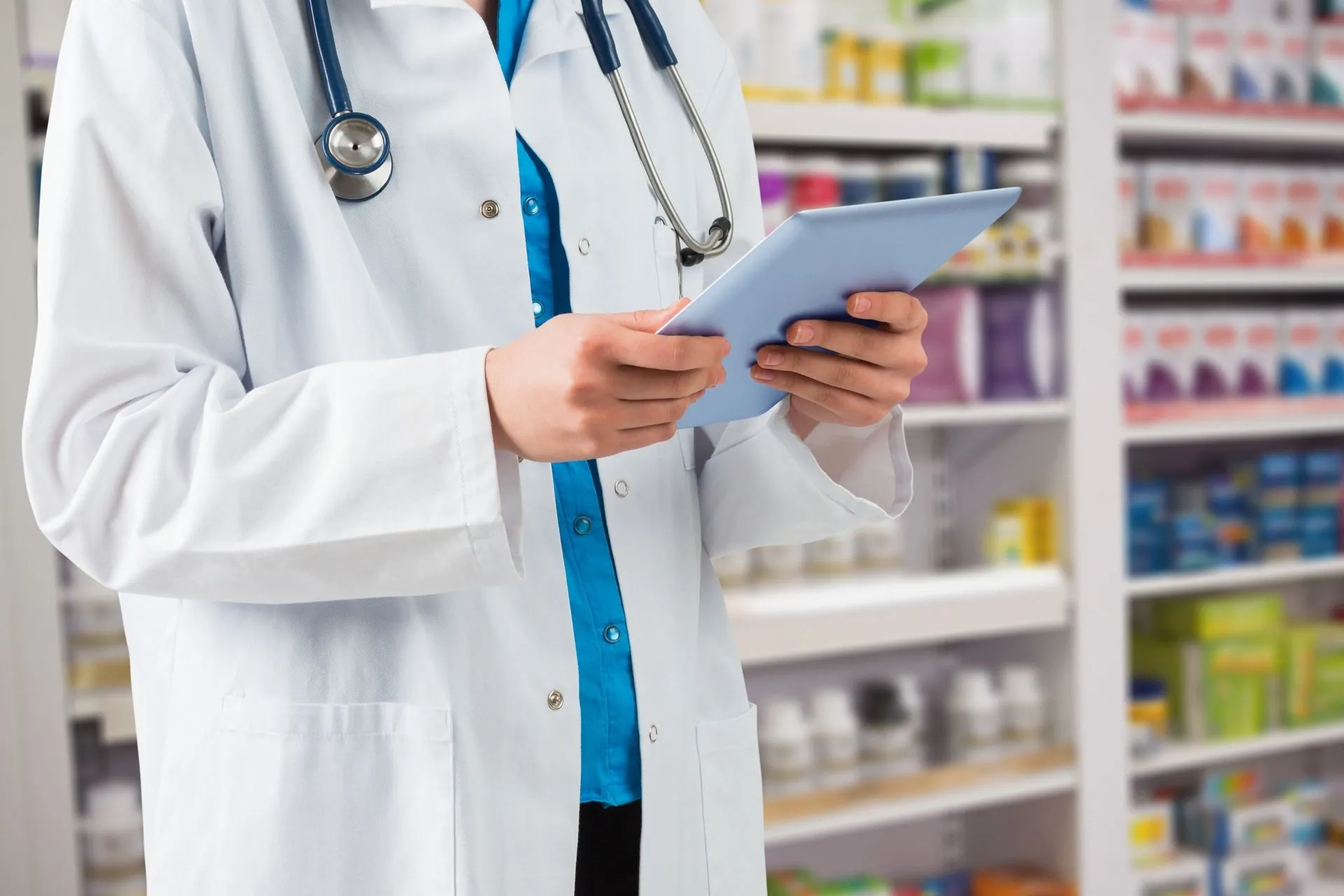 Pharmacist with a stethoscope reading a tablet in a pharmacy aisle