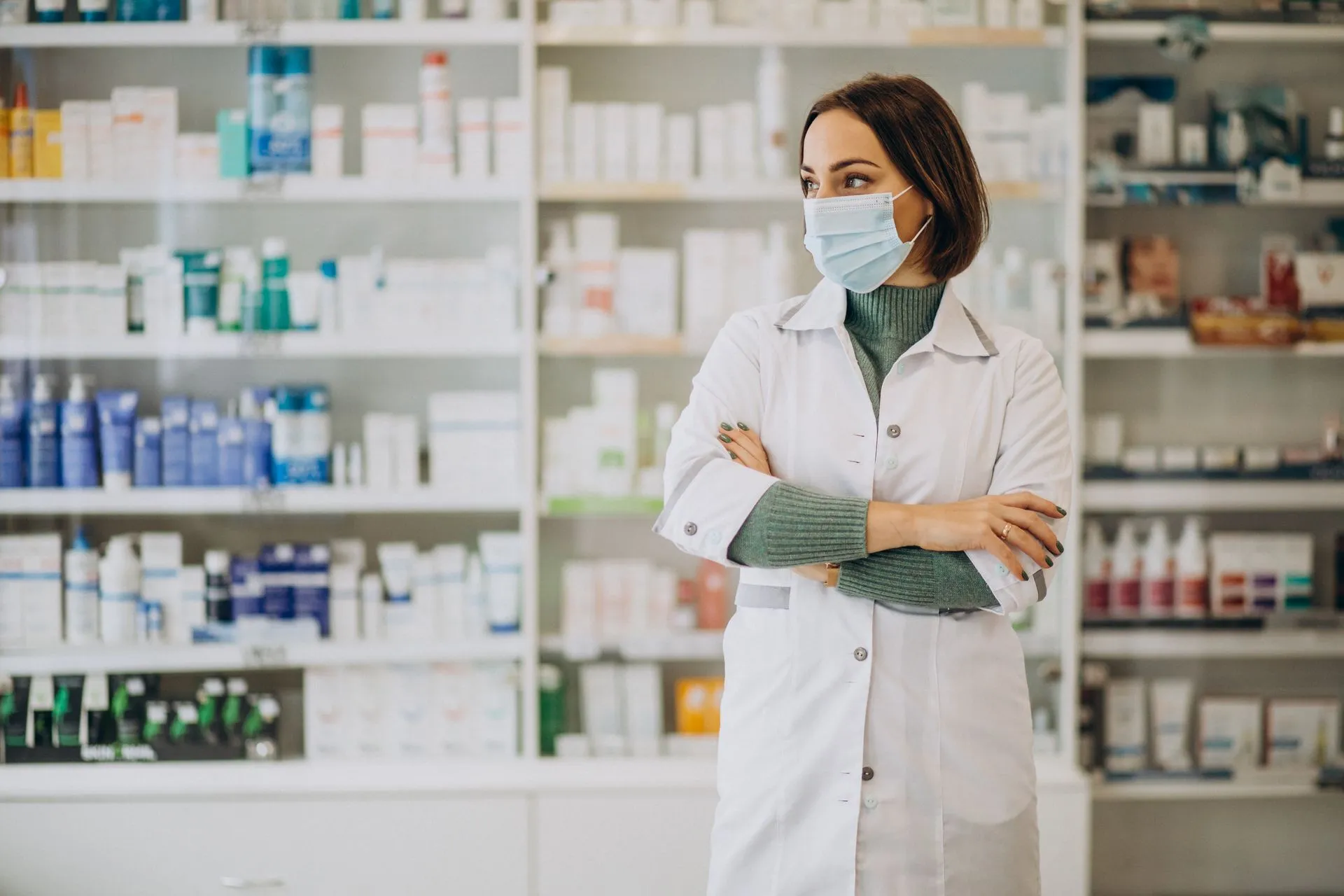 Pharmacist in a white coat and face mask standing in front of stocked pharmacy shelves